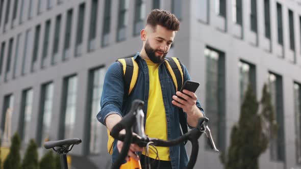 The Food Delivery Guy is Standing and Looking at the Smartphone