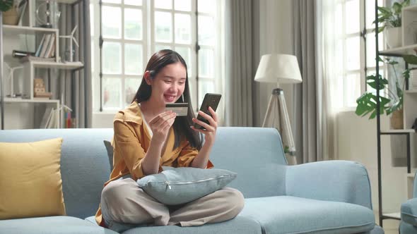 Asian Woman Sitting On Couch In Living Room Holding Credit Card And Using Smartphone Shopping Online