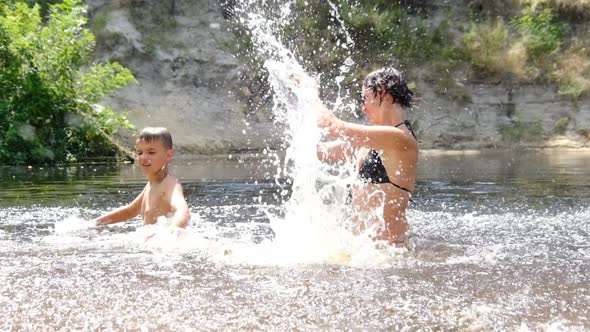 Happy Child and Mom are Splashing in a Shallow River Creating Waves Splashes