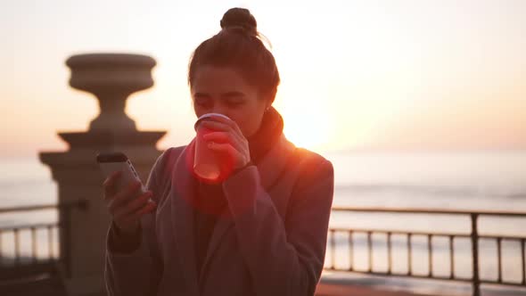 Young Business Girl Drinking Coffee and Surfing Internet Using Mobile Phone During Sunrise at Sea in