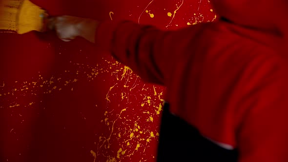 A Man in Hoodie Walking By the Red Wall and Drawing a Line with a Yellow Color Using a Brush