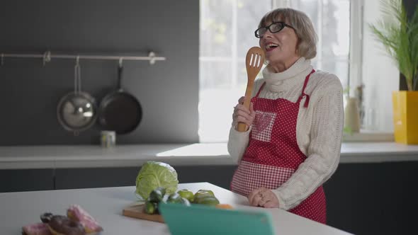 Cheerful Senior Woman Singing in Wooden Slotted Spoon Smiling Standing in Kitchen Indoors