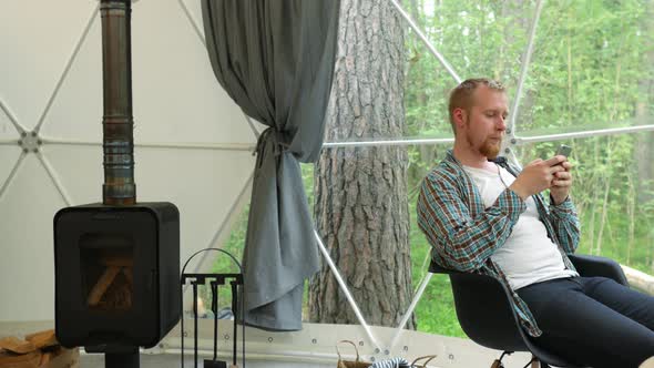 a man with a smartphone sits in a glamping tent overlooking the forest