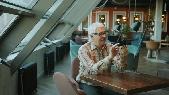 Senior Man Using Modern Smarthone Touching Screen Indoors at Table in Cafe