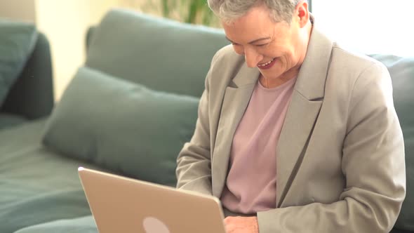 Smiling Grayhaired Lady in a Business Suit is Working with a Laptop at Home Alone While Sitting on