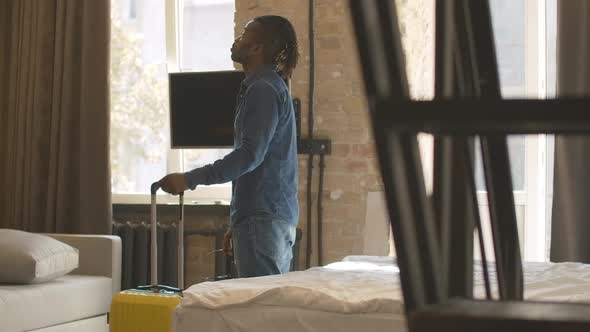 Joyful Relaxed African American Man with Luggage Looking Around in Hotel Room and Falling Back on