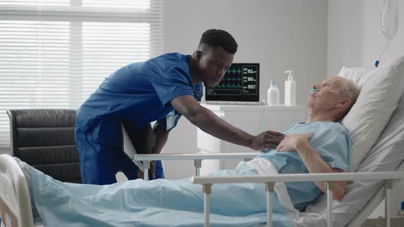 A Black Doctor of African Appearance is Talking to an Elderly Patient Lying on the Bed of the Clinic