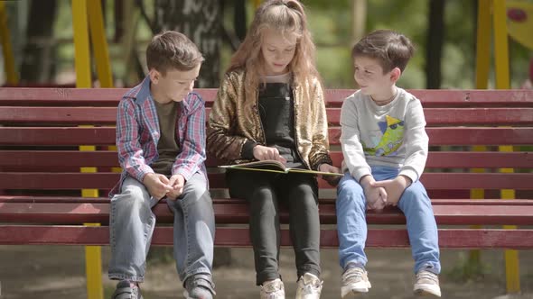 Cute Teen Girl Is Reading a Book To Two Younger Brothers While Sitting on a Bench Outdoors. Children