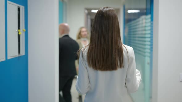 Back View of Slim Brunette Young Woman Walking in Office Hallway with Colleagues Passing