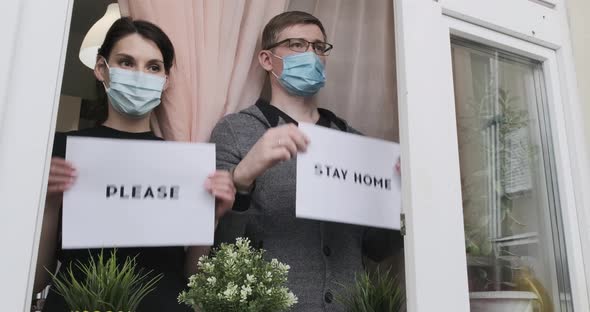 Young People in Medical Masked Show Poster That Says Stay Home in Window. Man and Woman Show Poster