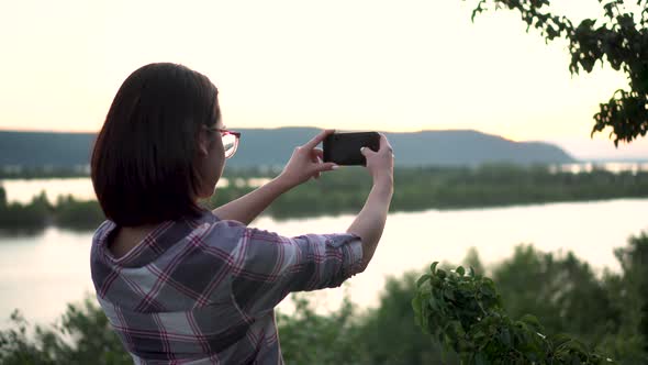 A Young Woman Stands on a Hill Against the Background of the River and Mountains and Photographs the