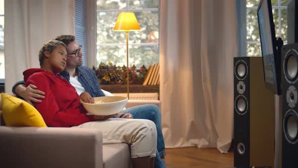 Side View of Multiethnic Young Couple Relaxing on Couch with Bowl of Popcorn and Watching Tv