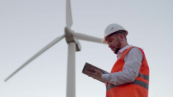 A Male Engineer in a Reflective Vest and White Helmet Looks at a Modern Tablet and Stretches Out His