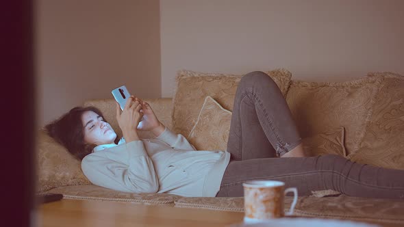 Teen Girl Lies at Night Near the TV on the Couch with a Mobile Phone