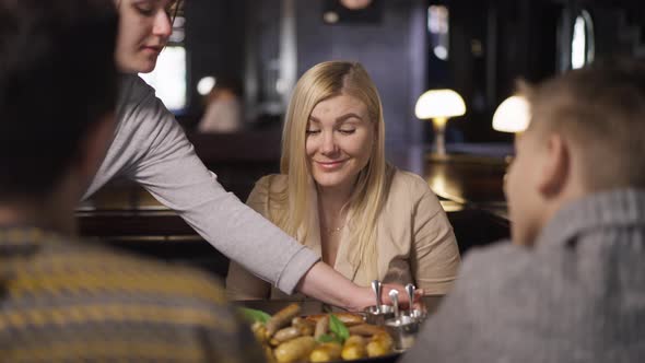 Confident Caucasian Woman Sitting in Restaurant with Husband and Son As Waiter Bringing Food in Slow