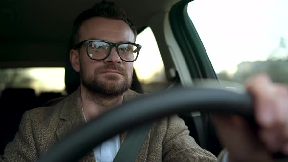 Satisfied Bearded Man in Glasses Driving a Car Down the Street in Sunny Weather