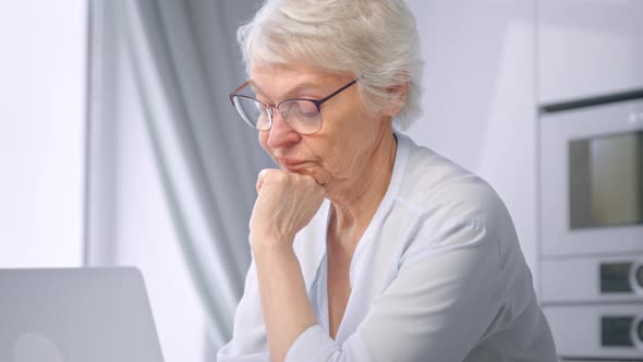 Concentrated old woman in glasses with short grey hairstyle types on laptop