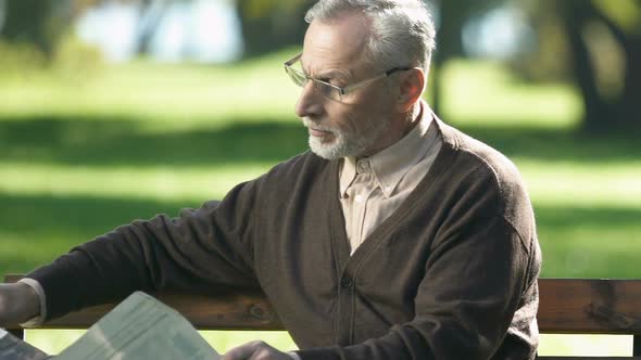 Retired Man in Glasses Reading Newspaper, Sitting on Park Bench in Morning