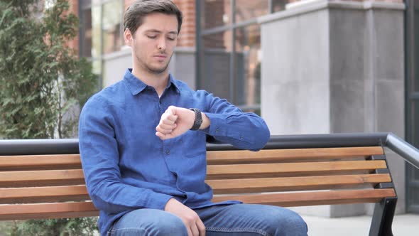 Waiting Young Man Watching Time on Watch, Sitting on Bench