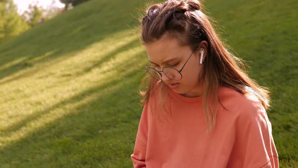 A Teen Girl is Sitting on the Green Grass with a Tablet and Wireless Headphones