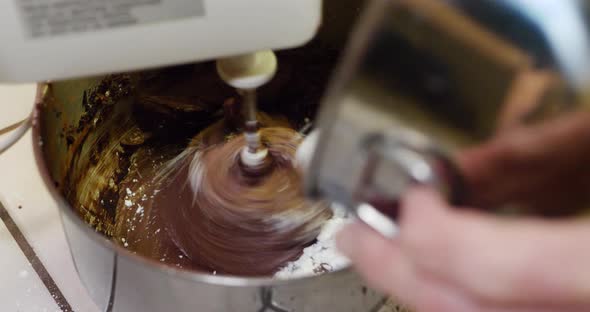 A chef in a kitchen using an electric mixer to make chocolate frosting for a cake in a metal mixing