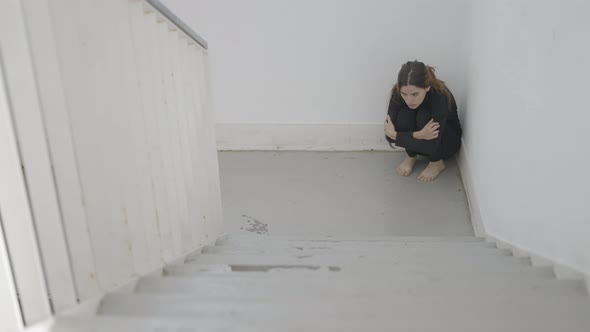 Young Scared Woman Sits in Corner of Stairwell with Arms Around Legs