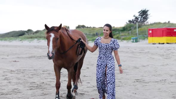 A beautiful girl with long hair in a blue dress walks her horse on the beach during the evening in D