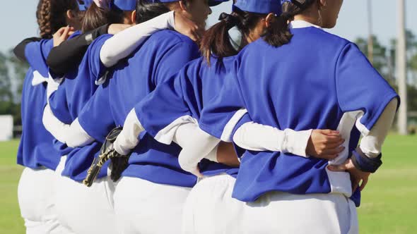 Happy diverse team of female baseball players standing on field with arms around each other