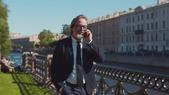 Portrait of Stylish Senior Entrepreneur Walking By River and Talking on Smartphone