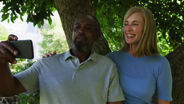 Diverse senior couple in garden taking selfie sitting under a tree and smiling