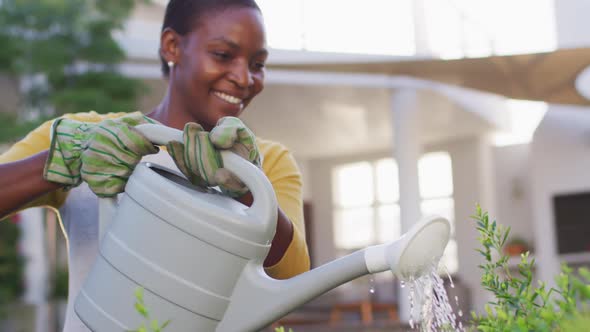 Happy african amercian woman wearing gloves watering flowers in garden