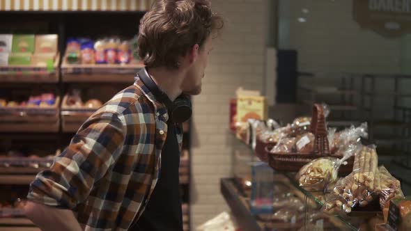 Young Guy with Headphones on Neck Choosing Tasty Cakes Choosing Pastries on Glass Showcase in Modern