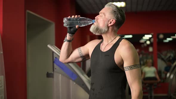 Gray-haired athletic man with tattoos drinks water in the gym between workouts