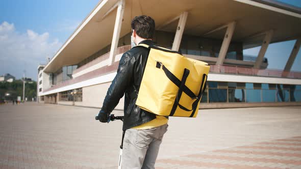 Back View of Delivery Man with Yellow Bagpack and Protective Mask Riding Electric Scooter with Food