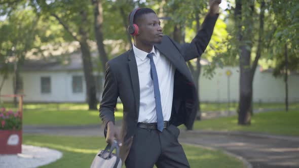 Portrait of Cheerful African American Businessman in Headphones Dancing in Sunny Park. Positive Man