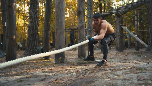 Sporty guy pulling a heavy tyre by a long rope. Man with naked torso workout in the forest gym
