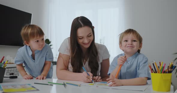 A Young Mother with Two Children Sitting at a White Table Draws Colored Pencils on Paper