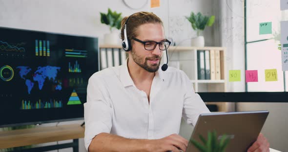 Man Sit at Workplace in Headset Looking at Laptop During Talking to Customer Using Remote Web Chat