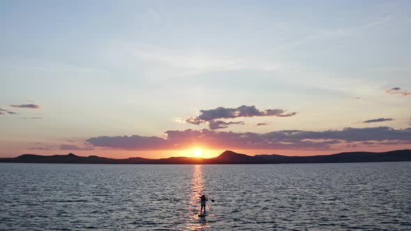 A Young Woman with Long Hair Stands on a Board Triumphantly Lifting Up an Oar at Sunset