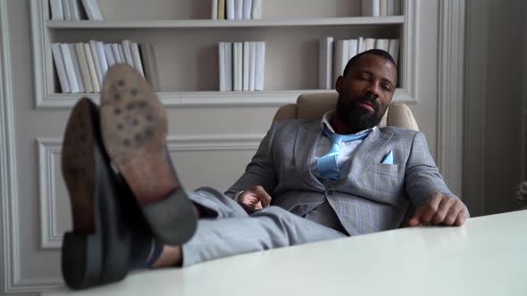 African-American with a Beard, Suit. The Businessman Is Sitting at the Workplace with His Feet on
