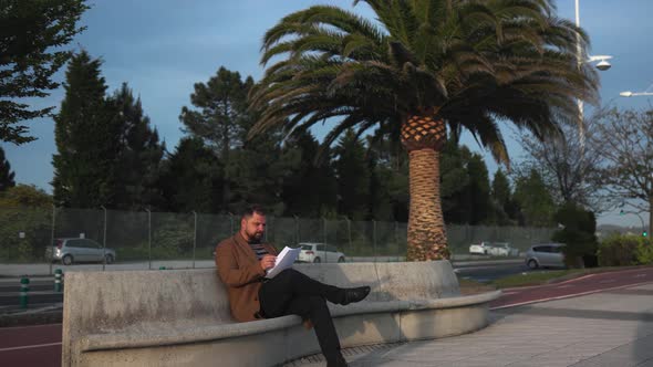 A Businessman in a Brown Coat Sits on a Bench and Looks at the Documents