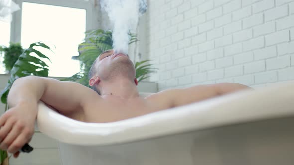 Sporty Naked Man Lies in Bathroom in Hotel or at Home with White Walls and Smoke Electronic