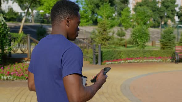 Afroamerican Man Uses Mobile Phone Holding White Cup with Coffee or Tea in Hand