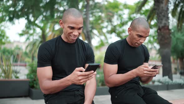 Two laughing bald African brothers looking at phone while sitting on the bench