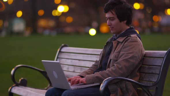 Young man sits on park bench using laptop computer