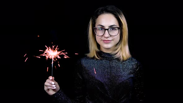 A Girl Holds a Sparkler in Hand. A Young Woman with Glasses Stands in a Shiny Evening Dress