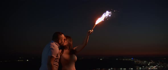 Young couple kissing while holding a firework at night, lights in the background