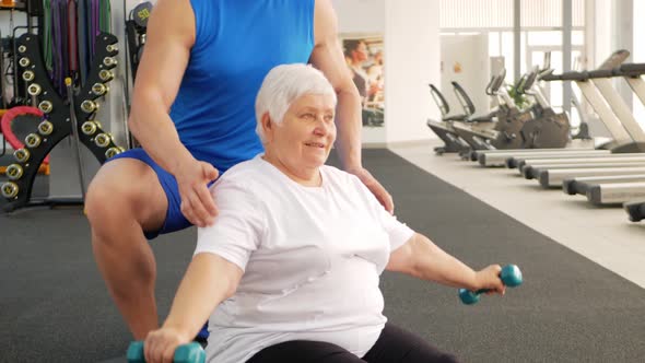 Pensioner Performs an Exercise with Dumbbells on Mat in Gym