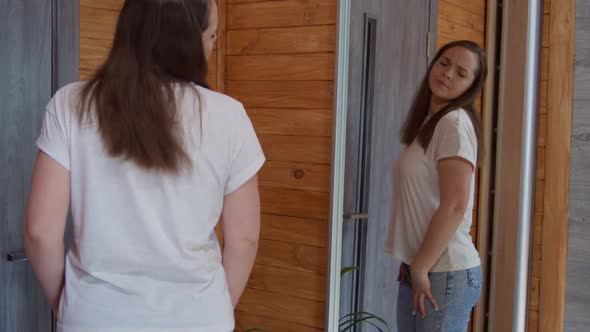 Young Average Woman in White Tshirt Examines Fold on Her Stomach Standing in Front of Mirror at Home