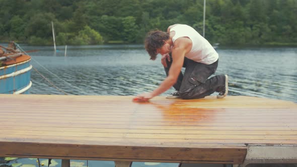 Youth walks into frame to start hand sanding roof of wooden boat planking.